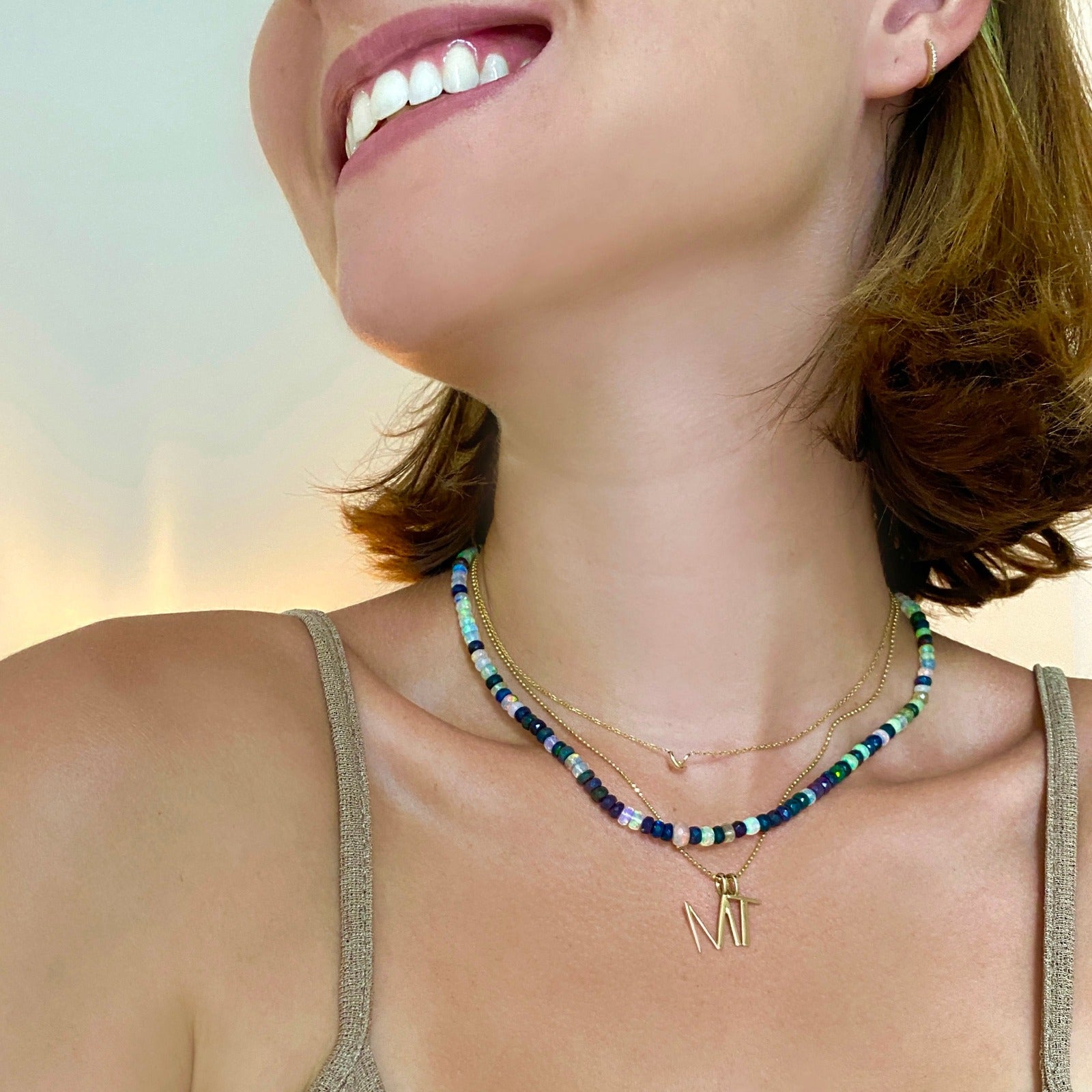Shimmering beaded necklace made of faceted opals in shades of fiery pastels, grey, teal, and black on a gold linking ovals clasp. Styled on a neck layered with a stitch necklace, bead chain necklace, M and T letter charms.