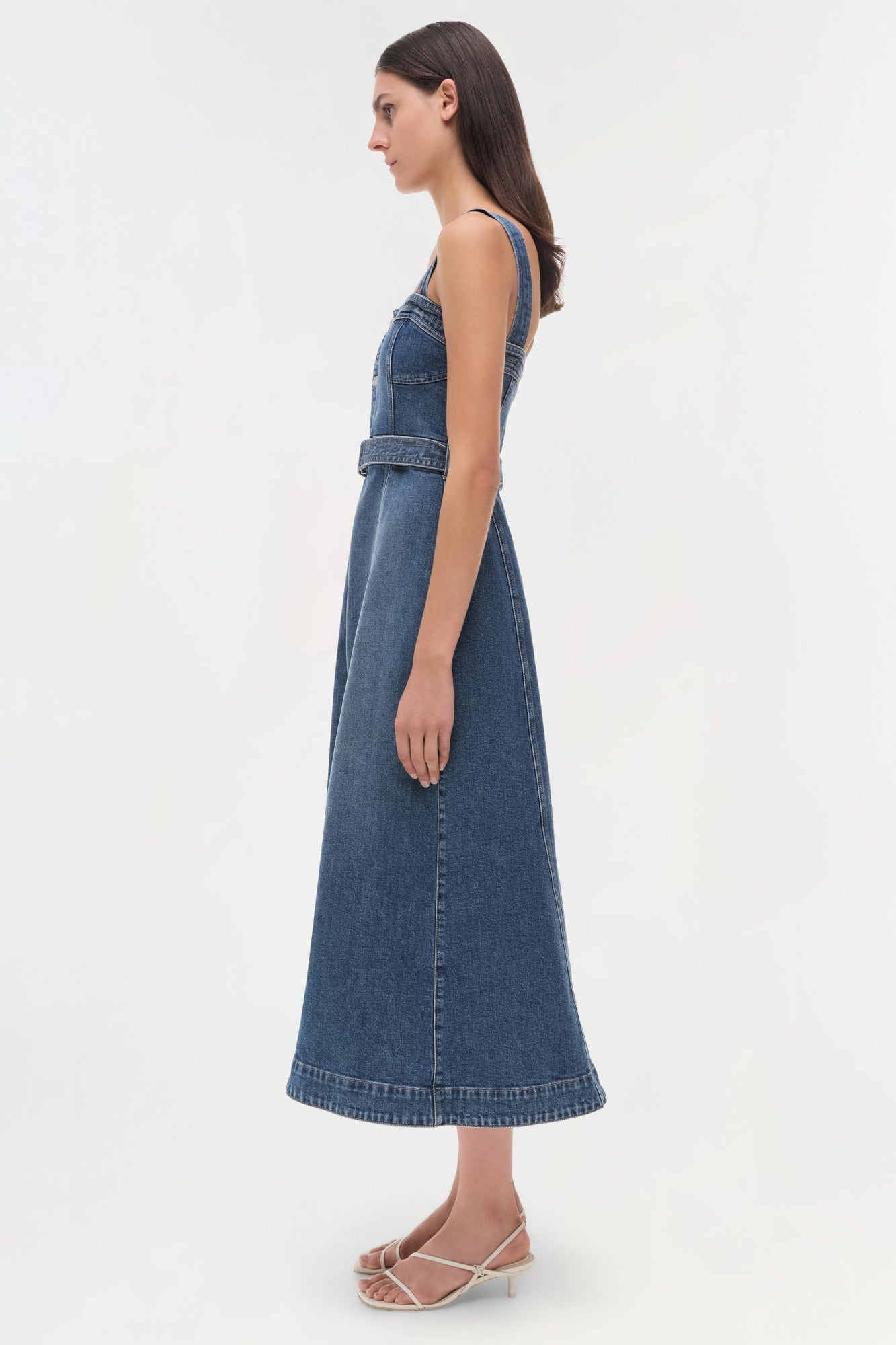 Woman wearing a blue denim sleeveless dress with a belted waist, shown from the side profile.