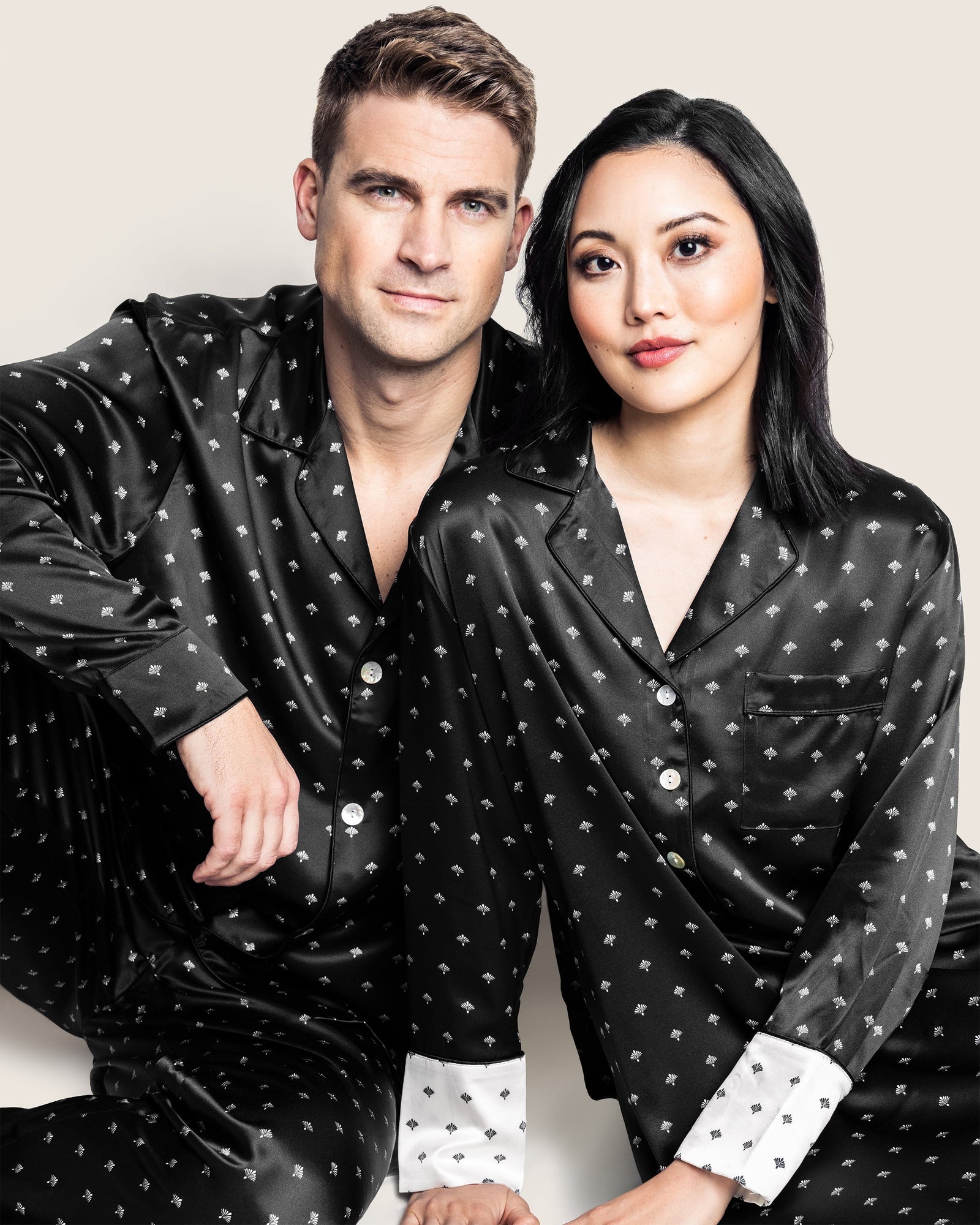 A man and woman sit together in matching Petite Plume Mens Silk Pajama Sets featuring a black Art Nouveau design. Their luxurious mulberry silk pajamas glow against the plain light backdrop as they gaze at the camera.