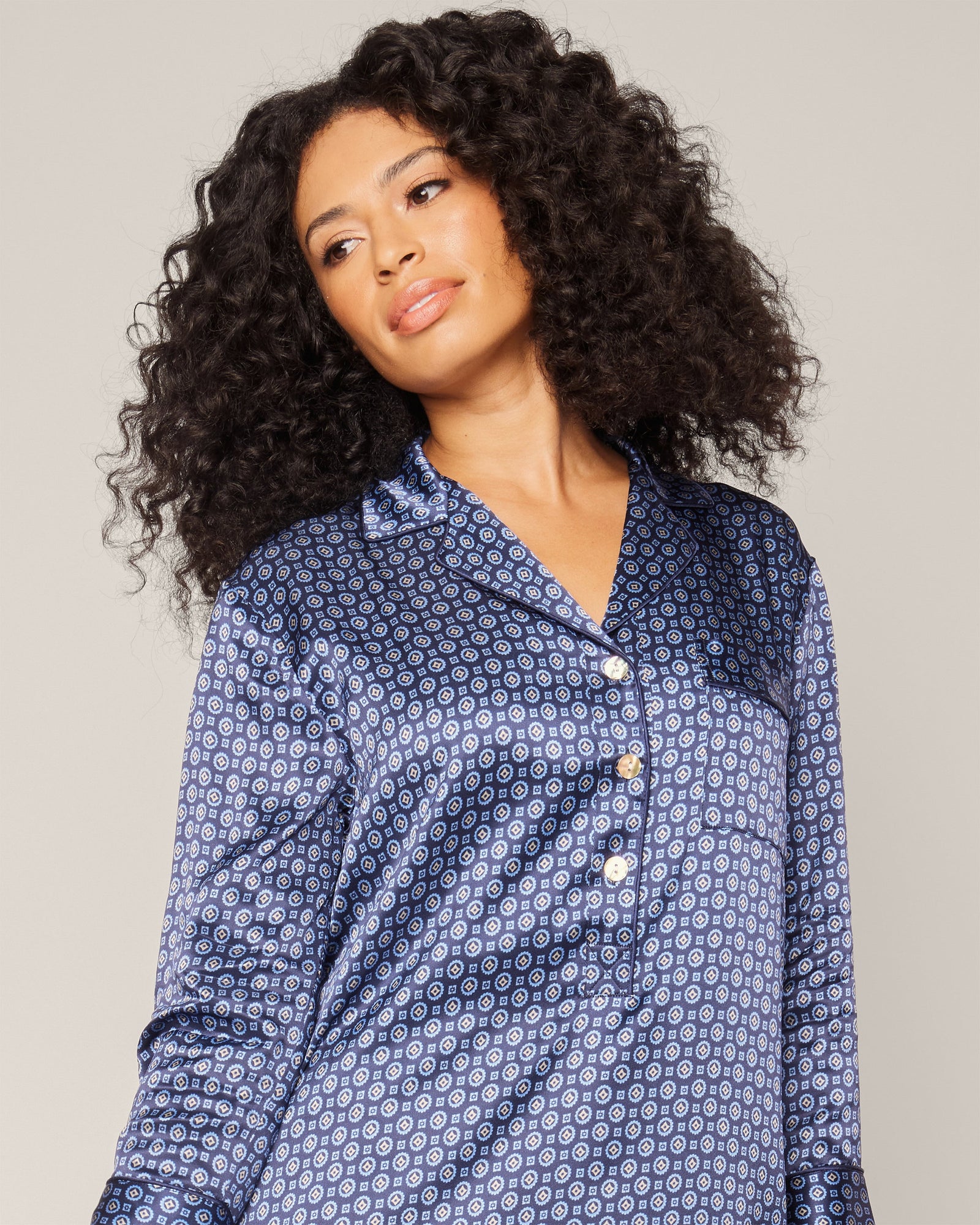 A person with long, curly hair wearing a Petite Plume Womens Silk Nightshirt in Midnight Foulard and a stylish neck scarf stands against a plain background, looking slightly to the side.