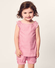A young child with curly brown hair is smiling, wearing Petite Plumes Girls Twill Amelie Short Set in Love Lines, featuring a pink and white striped top with ruffled sleeves and matching shorts. The background is a plain light color.