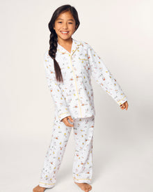 A young girl with long dark hair in a braid smiles barefoot, wearing the Kid's Twill Pajama Set in Easter Frolic, against a plain light background.