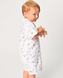 A young child with blonde hair wears the Baby's Twill Summer Romper in Easter Frolic—white with colorful animal prints and yellow trim—standing against a plain light background, holding two wooden blocks and looking to the side.