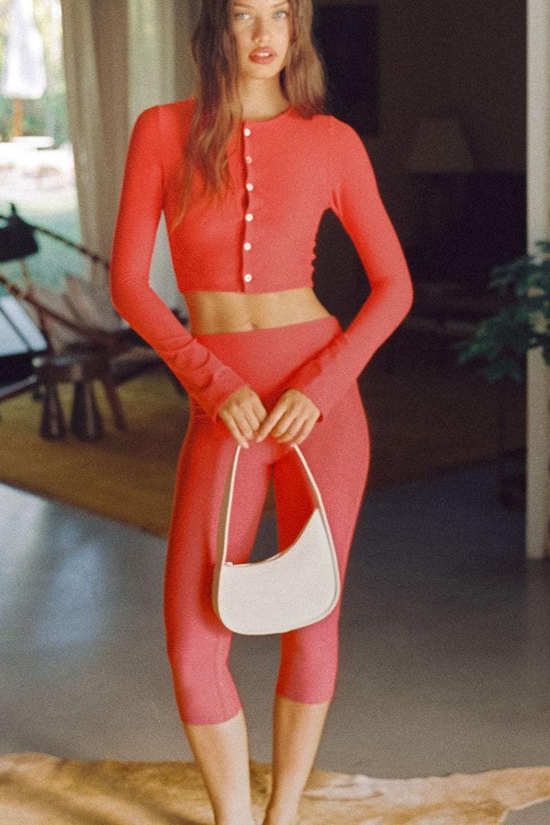 Woman wearing red crop top with button front and matching red capri leggings, styled with white shoulder bag.
