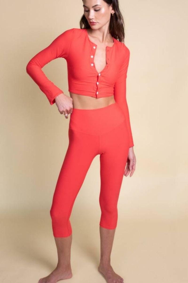 Woman wearing red capri leggings with matching cropped button-up long-sleeve top against neutral background.