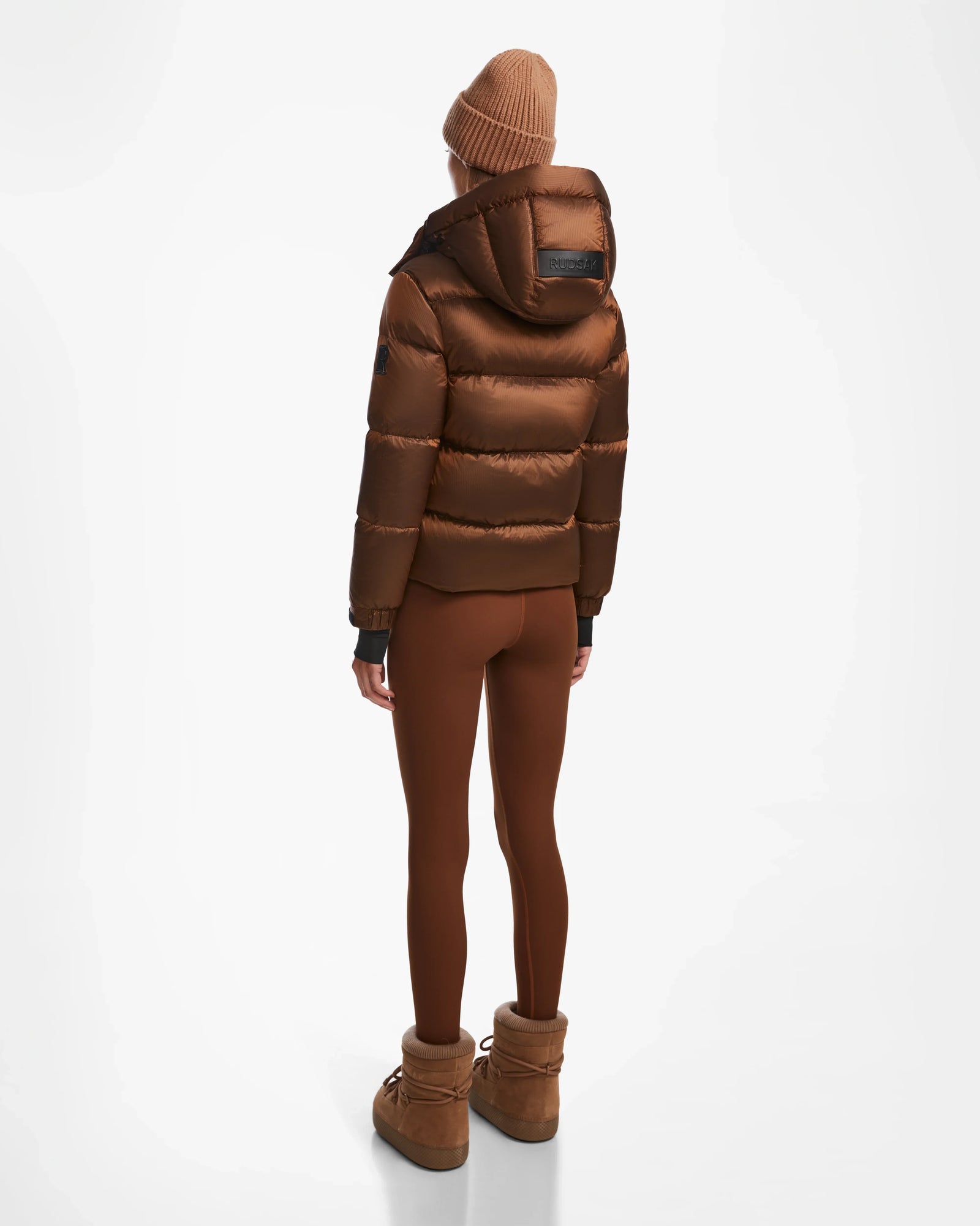 A person stands facing away, wearing the Rudsak ASHLYN S - 8124565 Antique Bronze womens down puffer jacket with a hood, paired with brown leggings, a matching beanie, and winter boots against a plain white background.