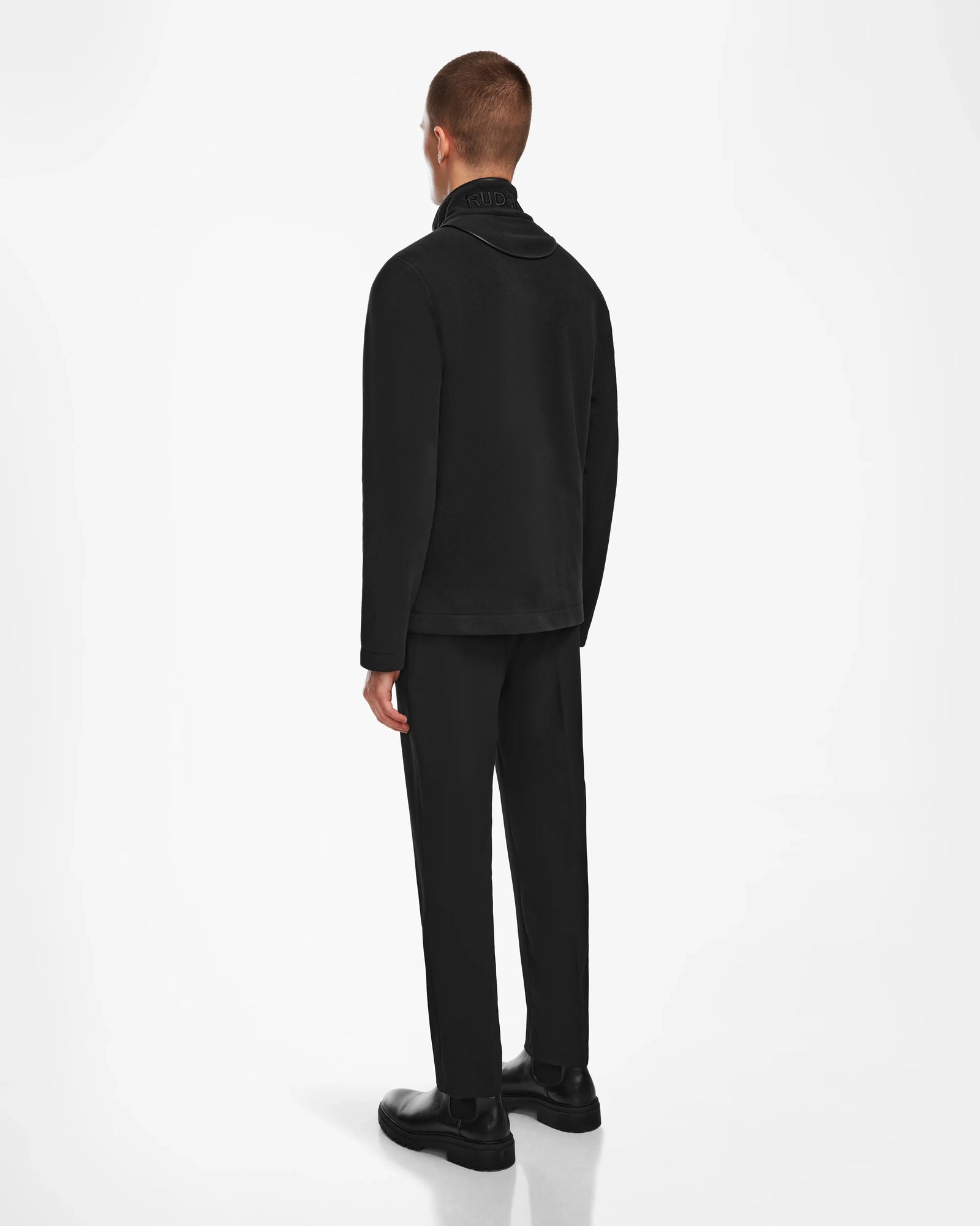 A person with short hair faces away from the camera, wearing Rudsaks BECK - 6524538 Black jacket with vegan leather trims, paired with black pants and shoes, set against a plain white background.