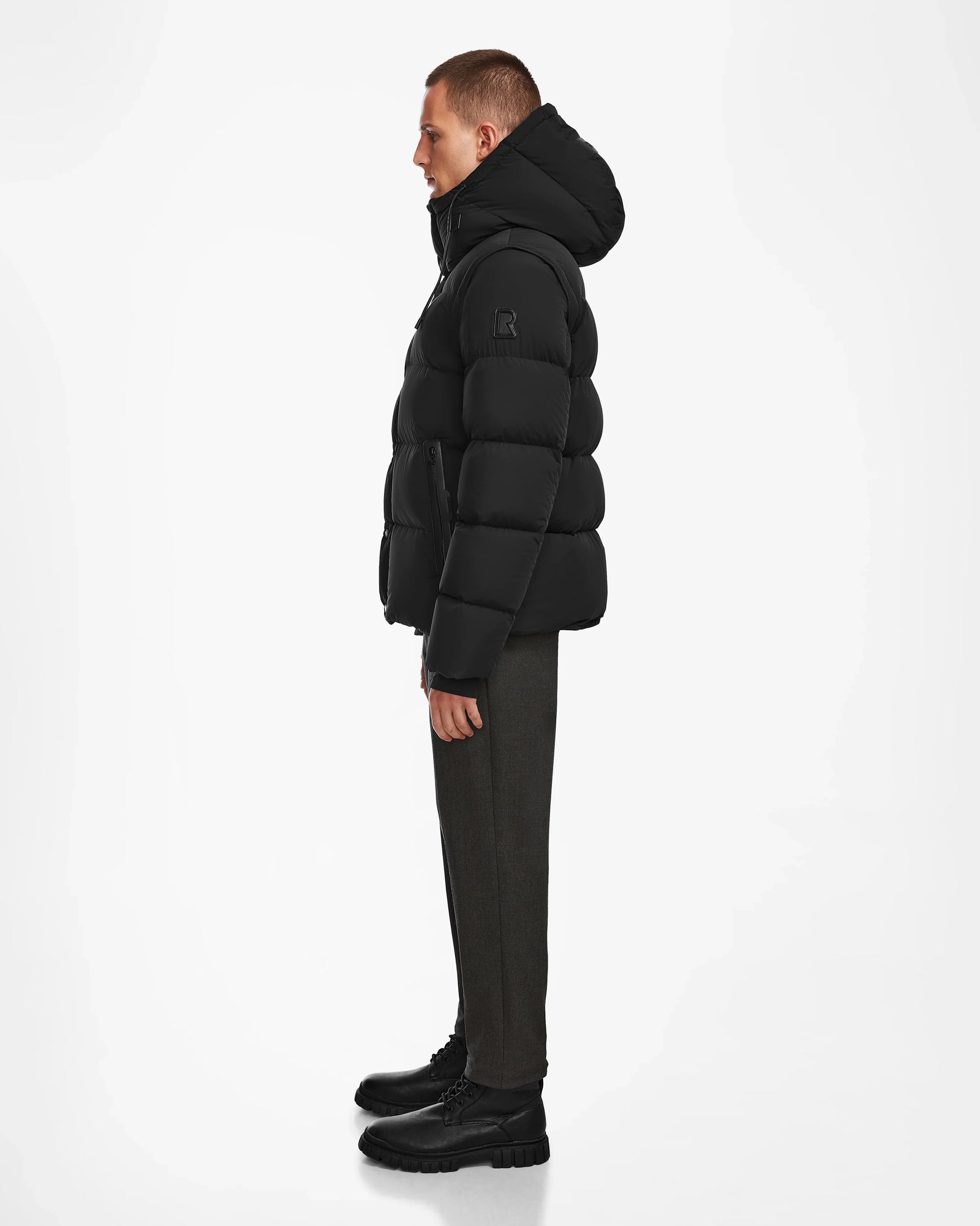 A person stands in profile against a white background, wearing the Rudsak BOBBY - 6124506 Black mens down puffer jacket with a hood, paired with dark pants and black boots.
