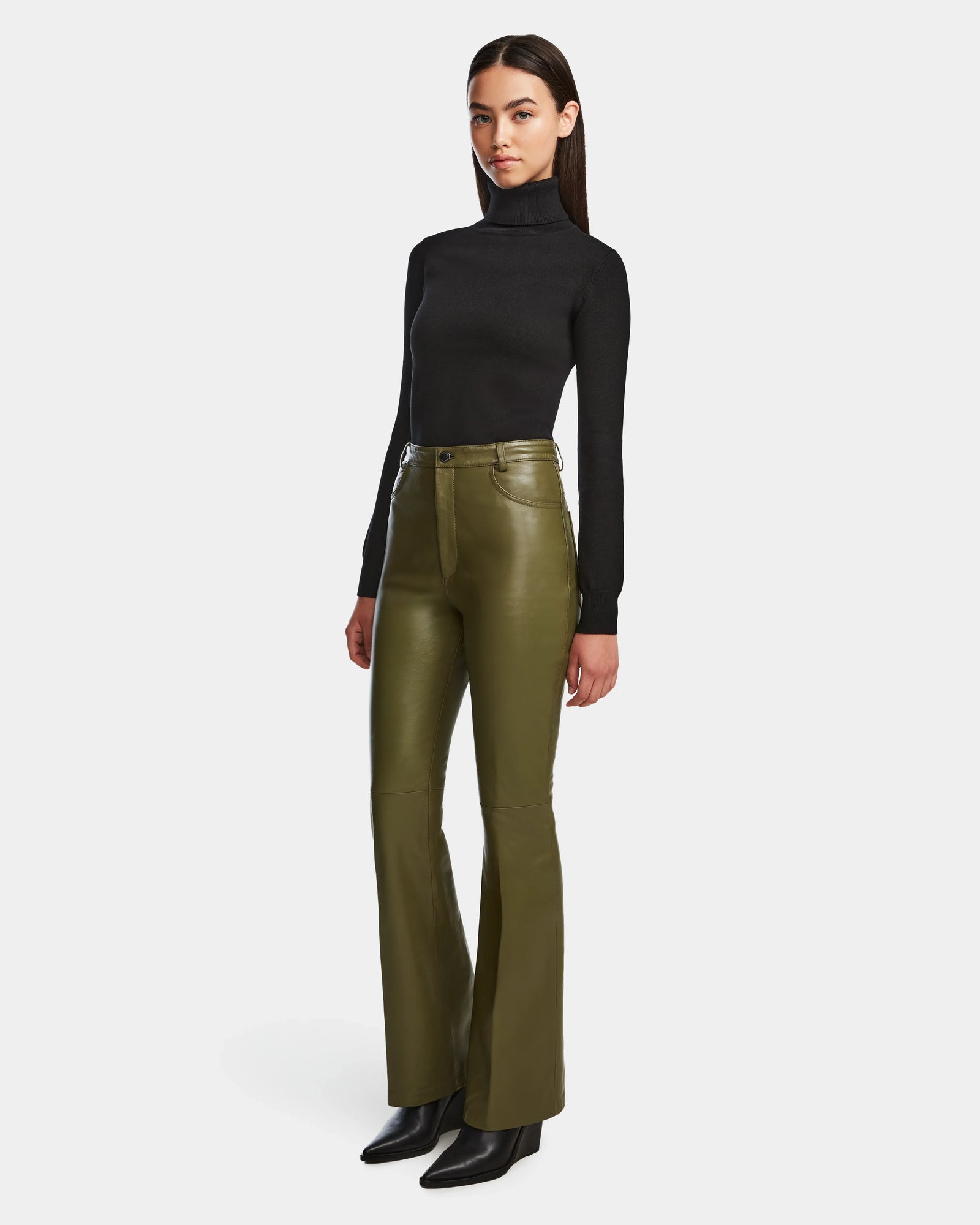 A woman with straight dark hair and a neutral expression stands against a white background wearing a fitted black turtleneck, Rudsak CARISSA - 8524545 Loden Green flare leather pants, and black heeled boots.
