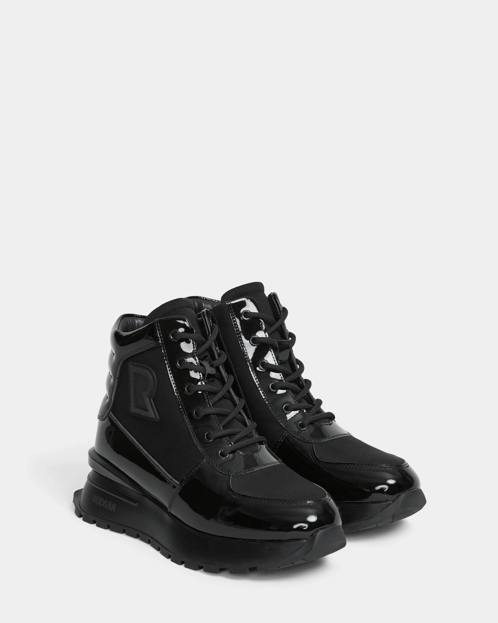 The CHELSA - L224505 Black by Rudsak are women’s sneaker boots with shiny patent leather accents, thick rubber soles, black laces, and side branding—offering a stylish upgrade to classic winter boots. Displayed on a plain white background.