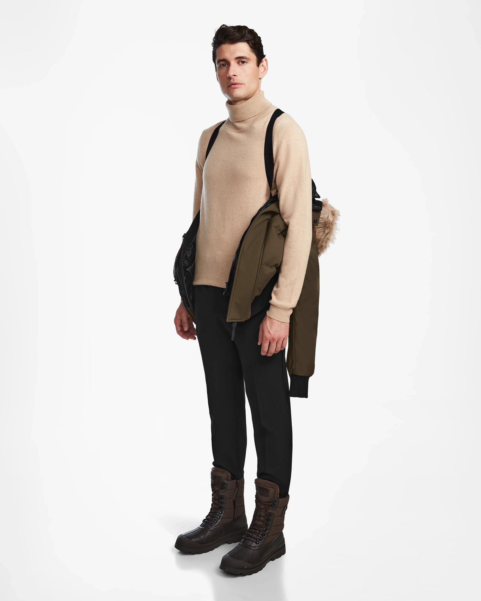 A man faces forward in a beige turtleneck, black pants, and brown boots, holding the Rudsak DALLAS - M122501 Army weather-resistant puffer jacket with a fur-lined hood over his shoulder against a white background.
