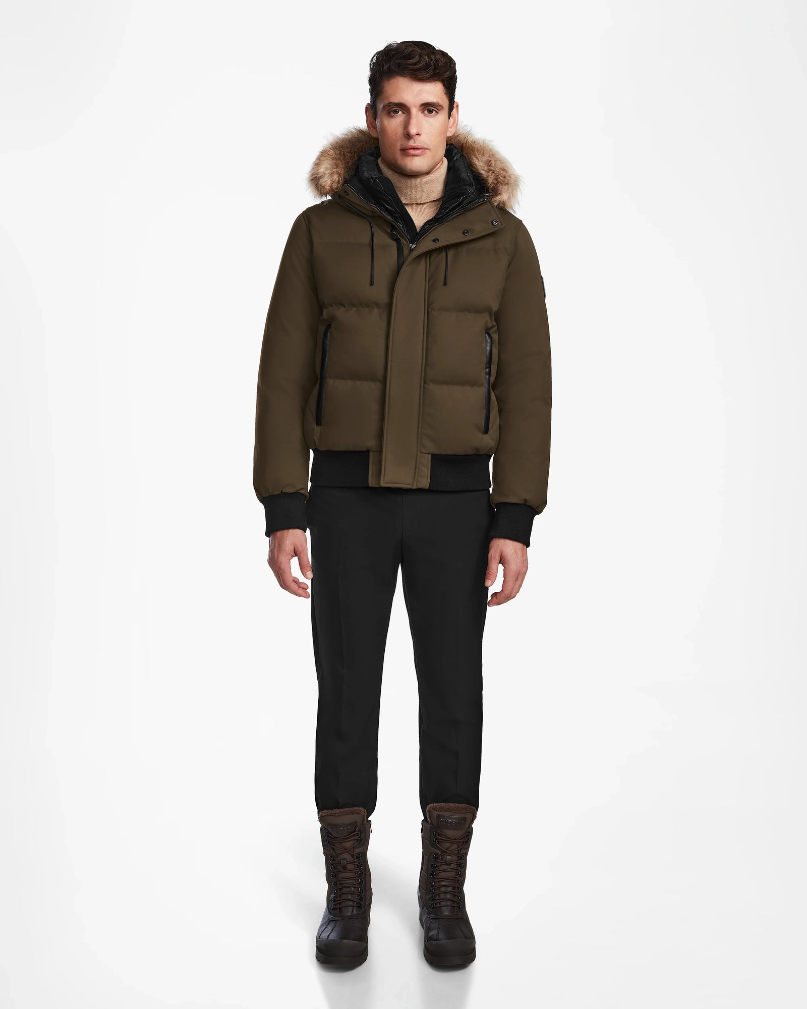 A man stands facing forward, wearing the Rudsak DALLAS - M122501 Army weather-resistant puffer jacket with a fur-trimmed hood, black pants, and brown lace-up boots against a plain white background.