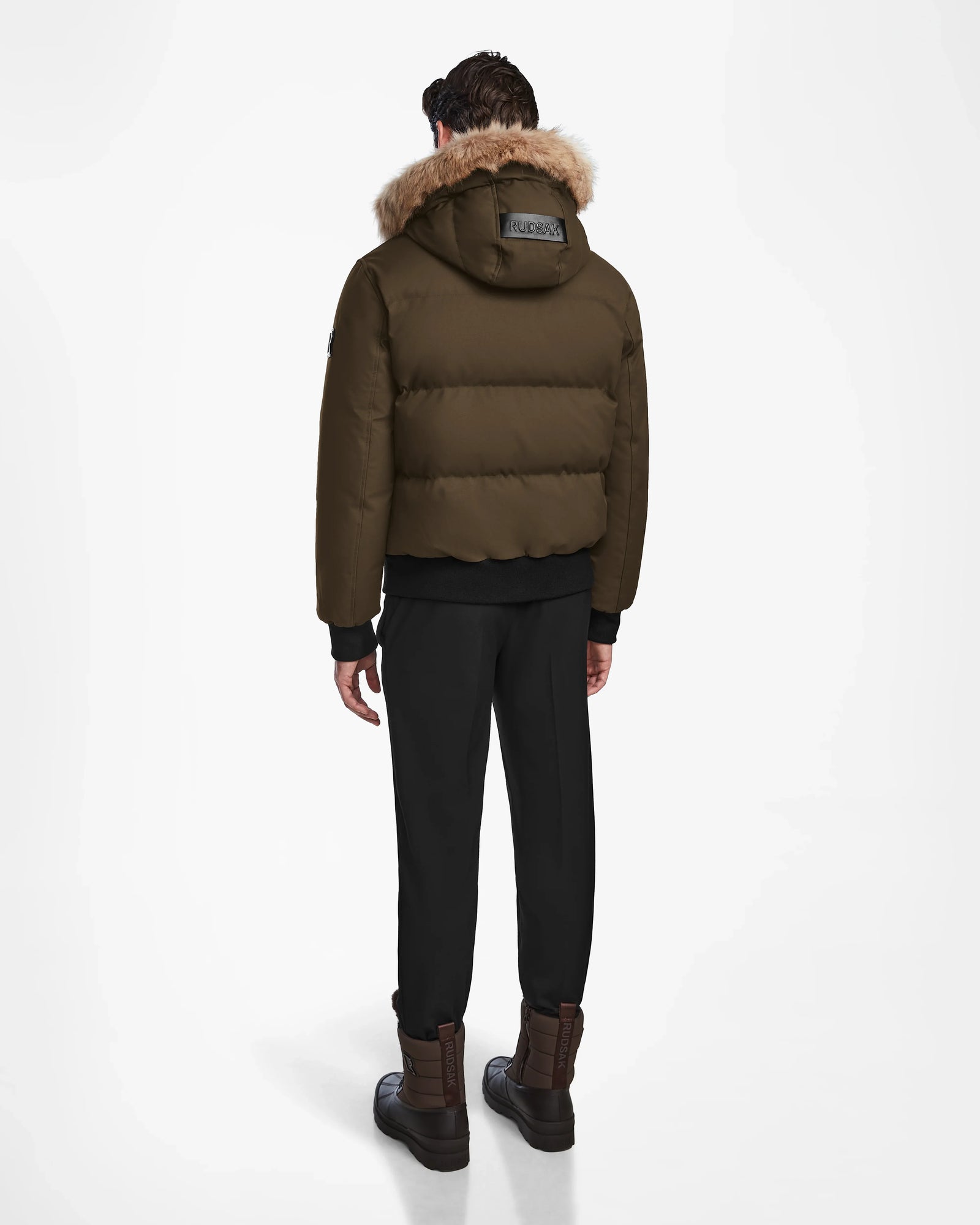A person faces away, wearing the Rudsak DALLAS - M122501 Army weather-resistant puffer with a fur-lined hood, black pants, and brown boots against a plain white background.