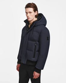 A young man with light skin and brown hair models the Rudsak DALLAS - M122501 Navy puffer, paired with black pants, standing against a white background and glancing toward the camera.