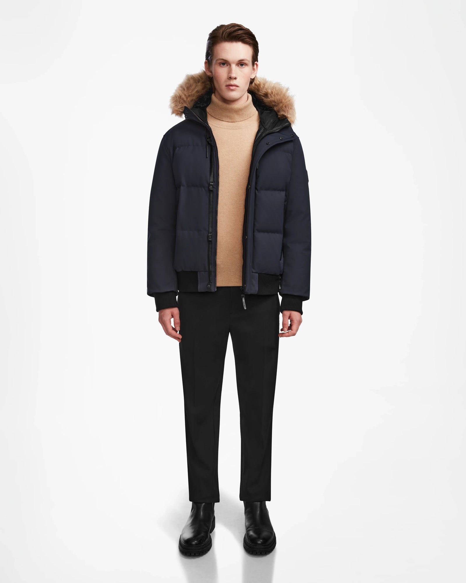 A person faces forward in the Rudsak DALLAS - M122501 Navy men's quilted down bomber with a fur-lined hood, paired with a beige turtleneck, black pants, and black boots against a white background.