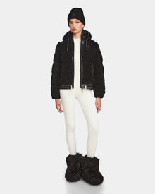 A person stands facing forward in the Rudsak DANI S - 8124537 Black bomber-style ski jacket, paired with a black beanie, white fitted outfit, and large black fuzzy boots against a plain light background.