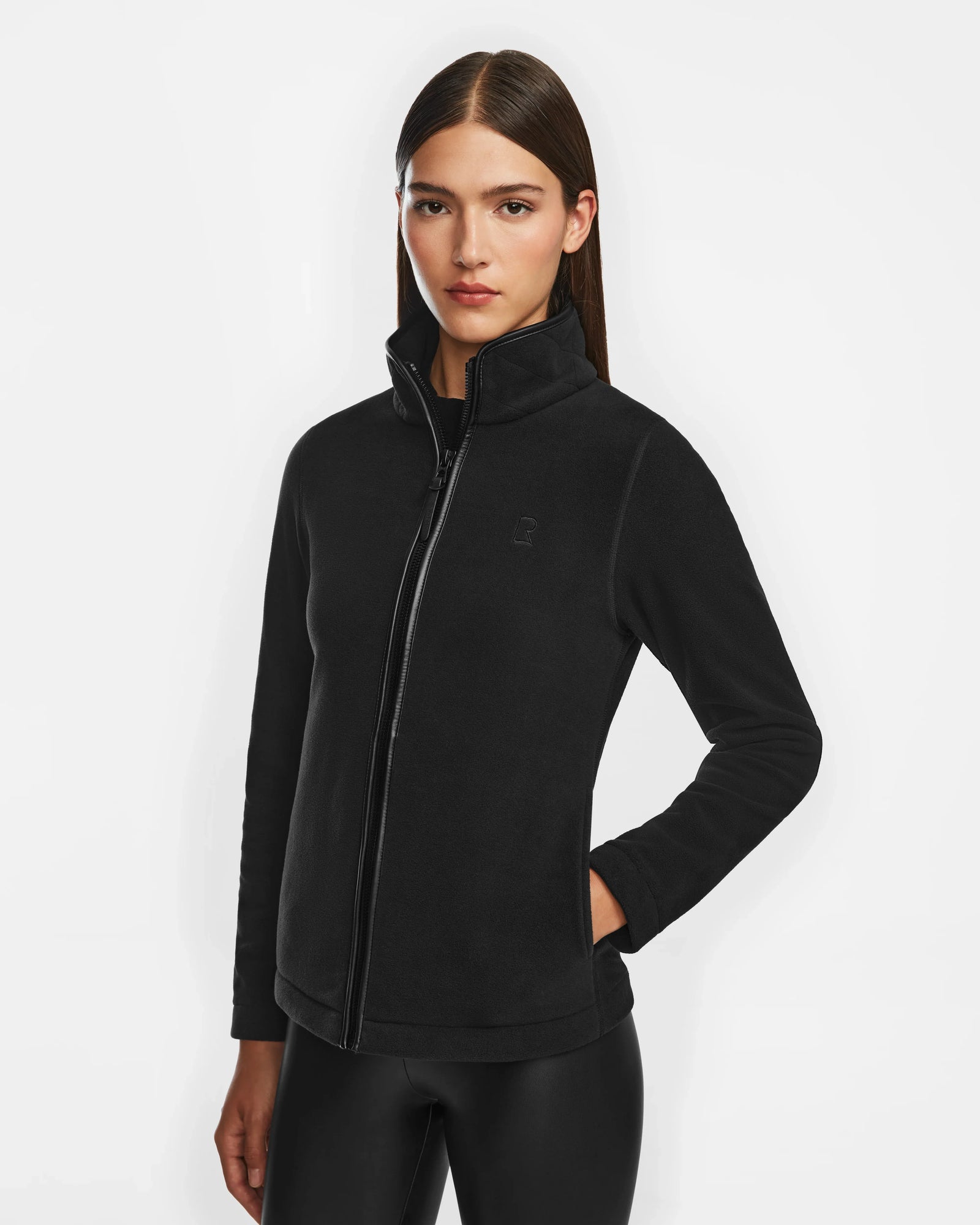 A woman with straight brown hair wears the Rudsak EDITH - 8524617 Black zip-up sweater with vegan leather trim and black leggings, standing against a white background and looking directly at the camera with a neutral expression.
