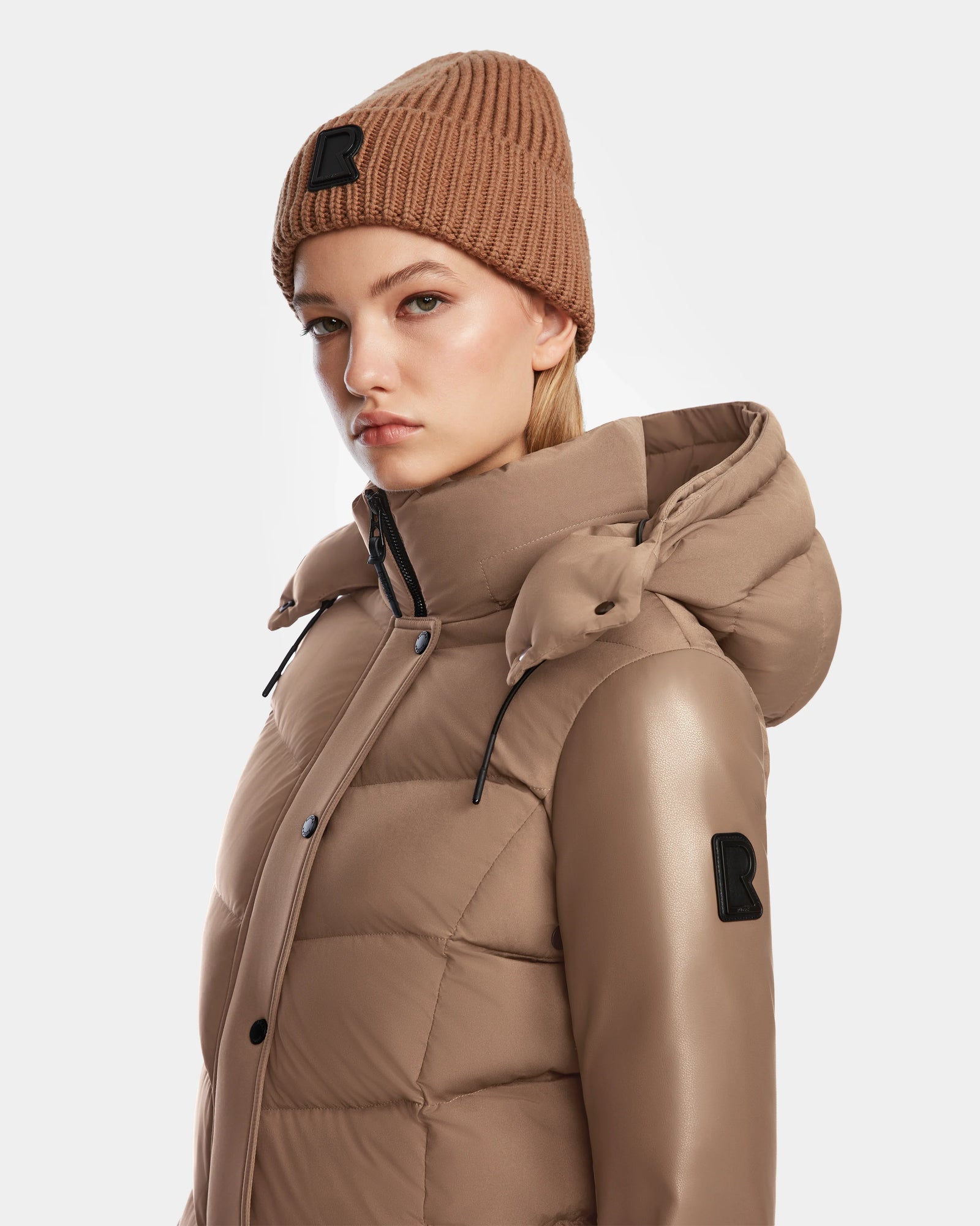 A woman wears the ELSA - L123535 Sand quilted down puffer jacket by Rudsak and a brown knit beanie, both with a black R patch, posing against a white background. The sustainable jacket offers eco-conscious style.