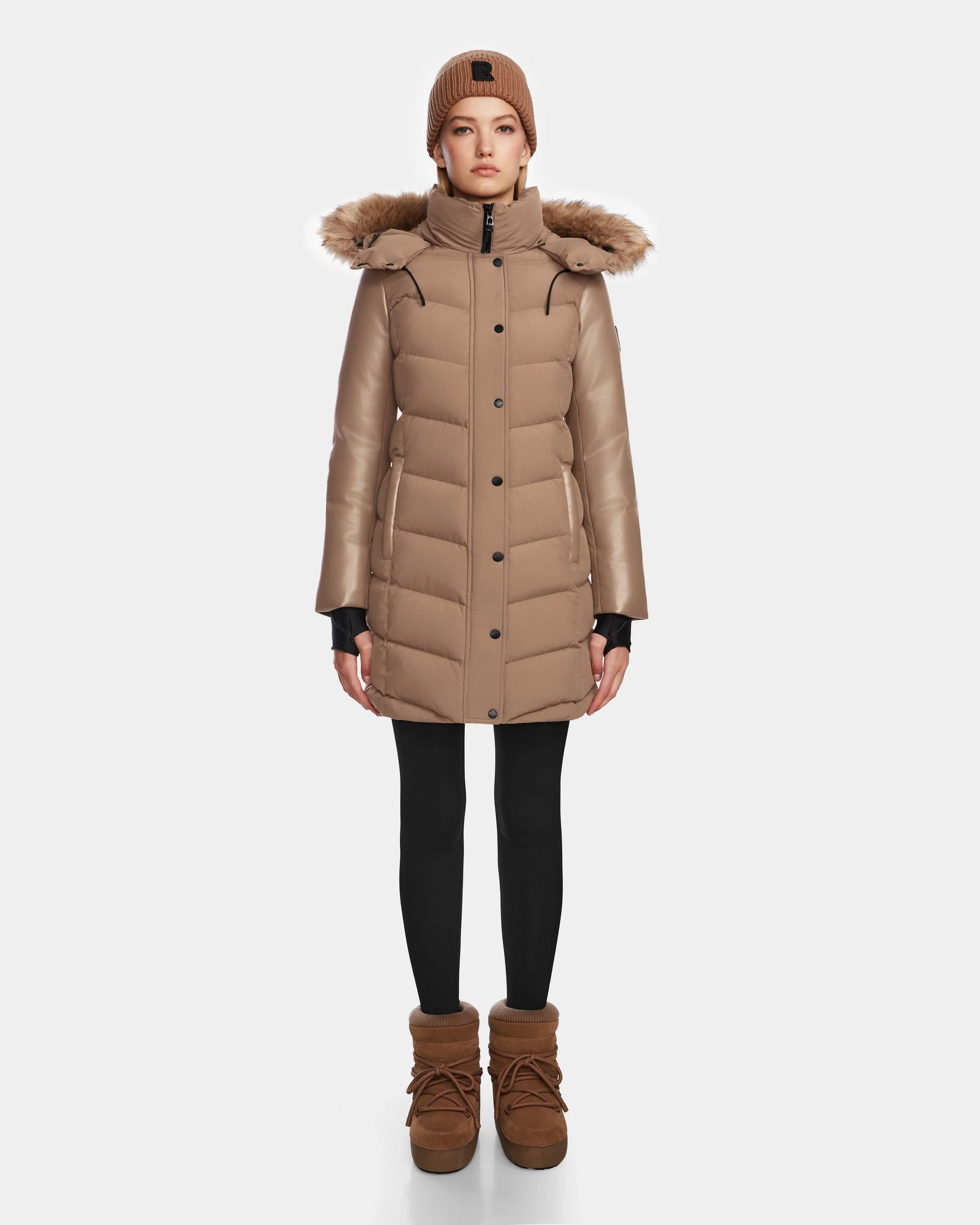A person stands facing forward, wearing the Rudsak ELSA - L123535 Sand women's quilted down puffer coat with a faux fur-trimmed hood, paired with a tan knit beanie, black leggings, and tan winter boots against a white background.
