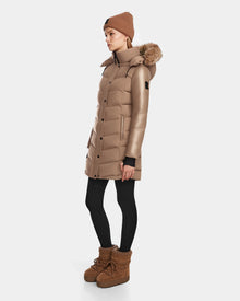 A woman stands sideways against a plain white background, wearing the Rudsak ELSA - L123535 Sand long brown quilted down puffer with a fur-trimmed hood, black leggings, brown winter boots, and a brown knit beanie.