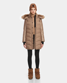 A person stands facing forward, wearing the Rudsak ELSA - L123535 Sand women’s quilted down puffer coat with fur-trimmed hood, a matching knit beanie, tan sweater, black leggings, and tan boots against a plain white background.