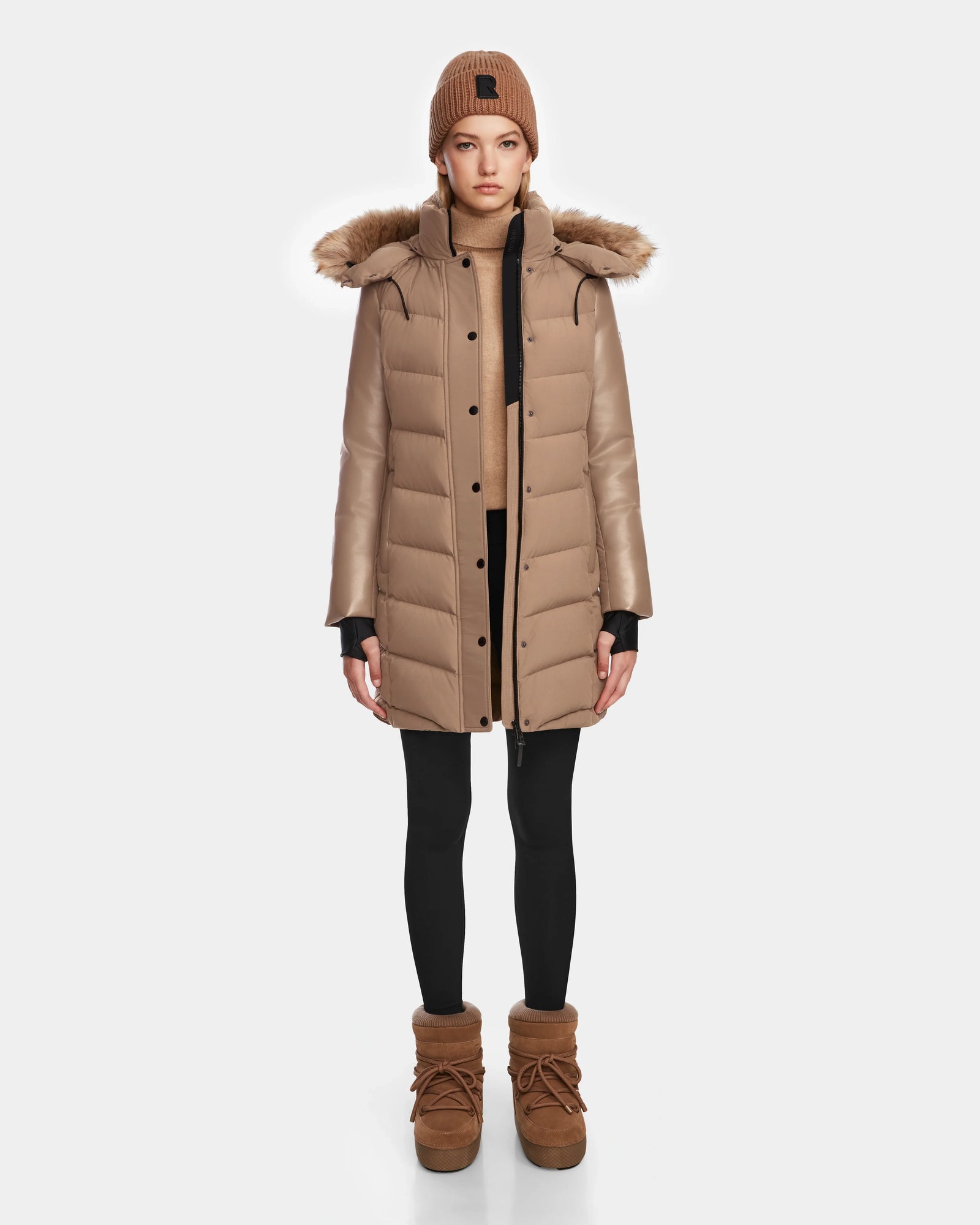 A person stands facing forward, wearing the Rudsak ELSA - L123535 Sand women’s quilted down puffer coat with fur-trimmed hood, a matching knit beanie, tan sweater, black leggings, and tan boots against a plain white background.