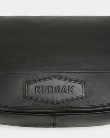 Close-up of the GRAYSON L - 8324525 black leather crossbody bag featuring a stitched rectangular patch embossed with the Rudsak brand name.