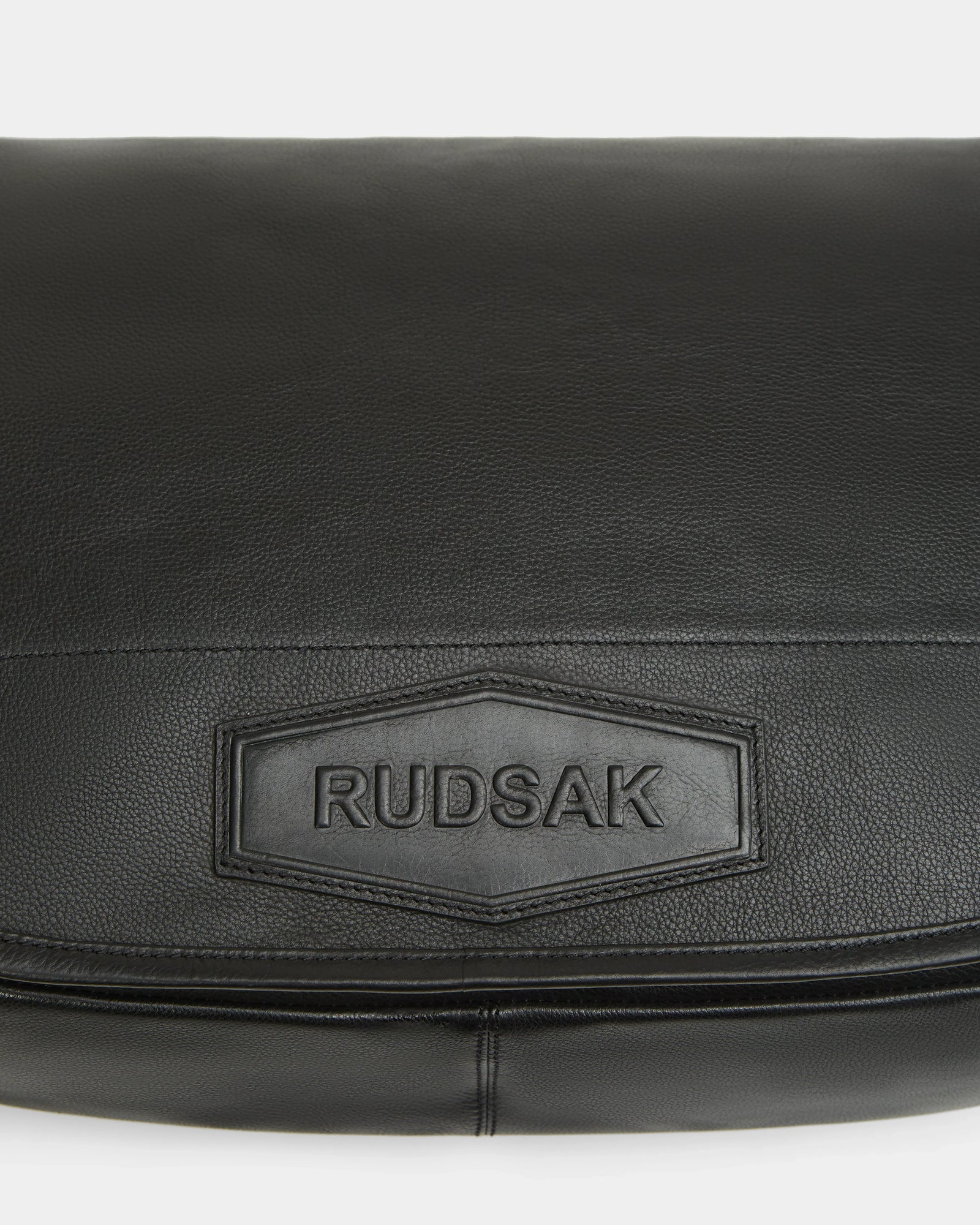 Close-up of the GRAYSON L - 8324525 black leather crossbody bag featuring a stitched rectangular patch embossed with the Rudsak brand name.