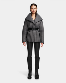 A woman faces forward on a plain white background, wearing the Rudsak INES T - 8124625 Grey quilted jacket with a wide collar, black belt, black leggings, and knee-high black boots.