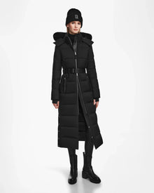 A woman faces forward against a plain white background, modeling the Rudsak IZABELLA - L124541 Black quilted maxi coat with a belt, paired with black boots, leggings, a beanie, and a dark turtleneck—highlighting this winter essential.