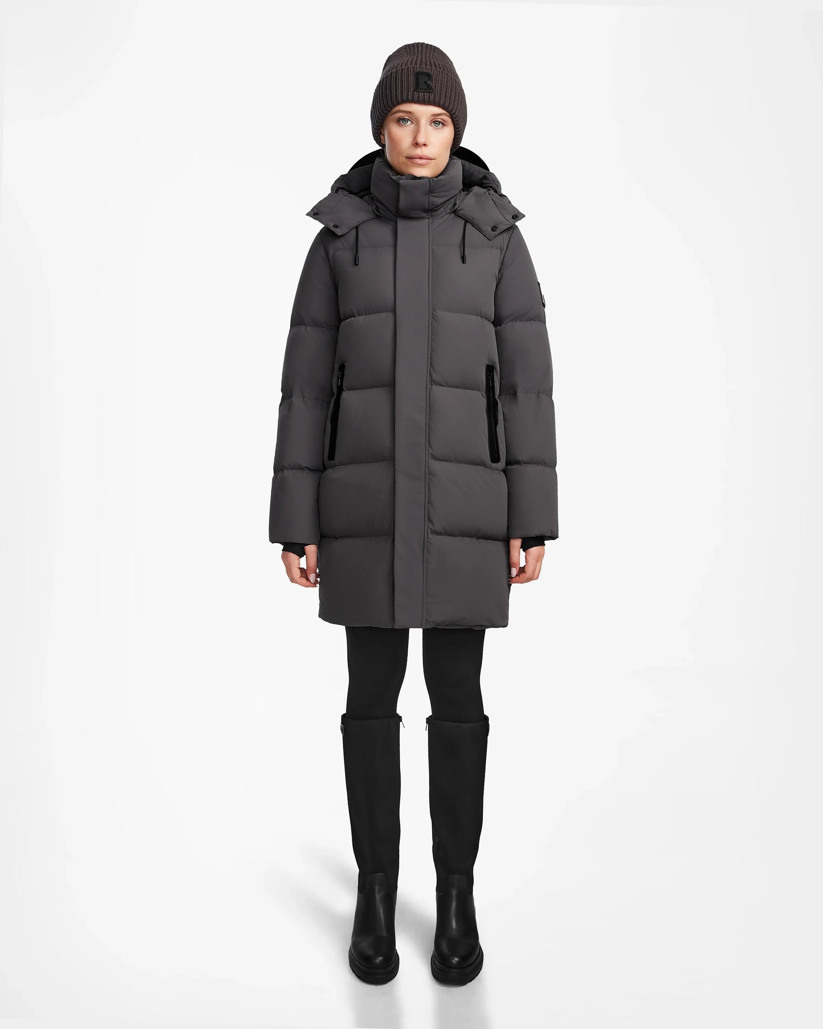 A person faces forward in a long Rudsak JOELLE - 8124729 Grey RDS-certified down puffer coat, with black leggings, boots, and a dark knit beanie. The background is plain white.