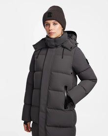 A woman in the Rudsak JOELLE - 8124729 Grey dark gray quilted down coat with a high collar and matching knit beanie stands with one hand in her pocket against a plain white background.