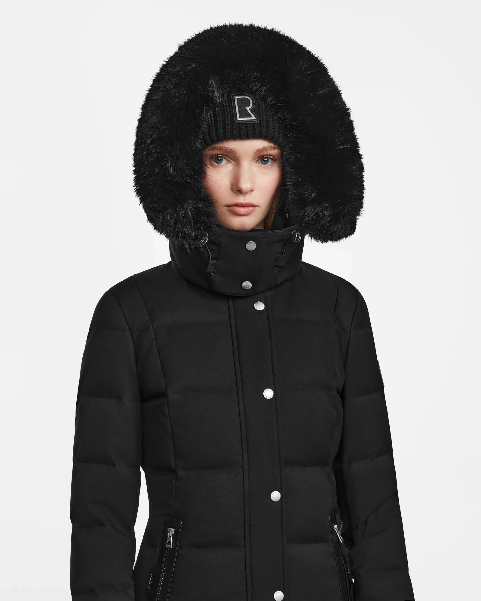 A woman wearing the Rudsak KEHLANI - L124540 black quilted down coat with a large faux fur-trimmed hood and a black beanie stands against a plain white background.