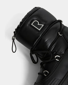 Close-up of the Rudsak KIKA - L224506 Black women’s winter boot featuring padded black material, black laces, silver eyelets, a bold metallic R logo on the upper cuff, and a chunky rubber sole, shown against a plain white background.