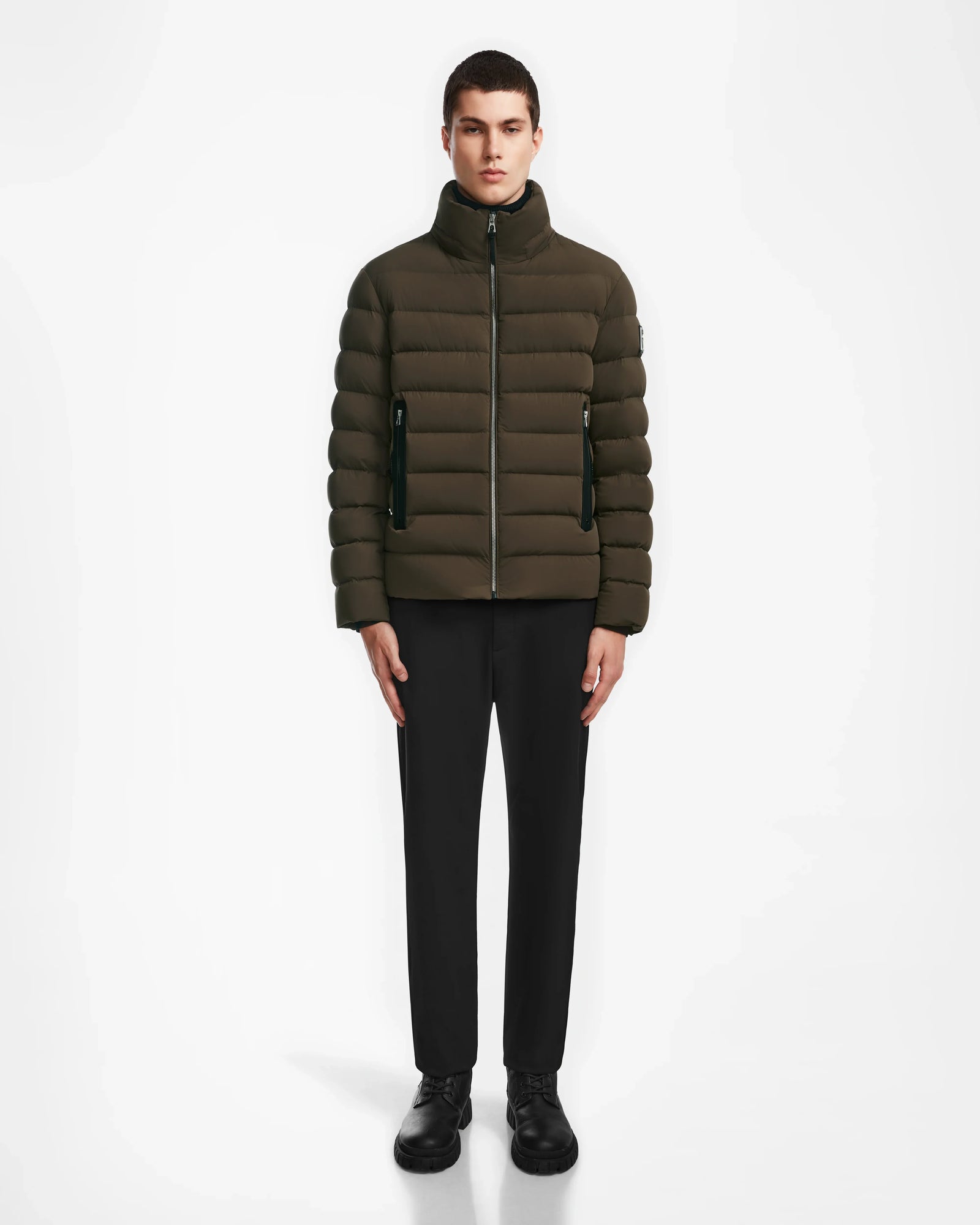 A man stands facing forward, wearing the Rudsak NAVAN - 6124551 Army light down puffer with a high collar, paired with black pants and black shoes against a plain white background.
