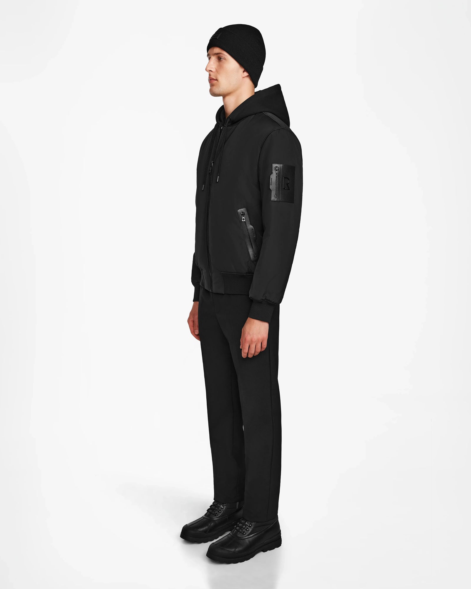 A man stands facing slightly left against a white background, wearing the Rudsak NEVEN - M124520 Black vegan leather men’s bomber jacket with zippered pockets, paired with black pants and shoes.