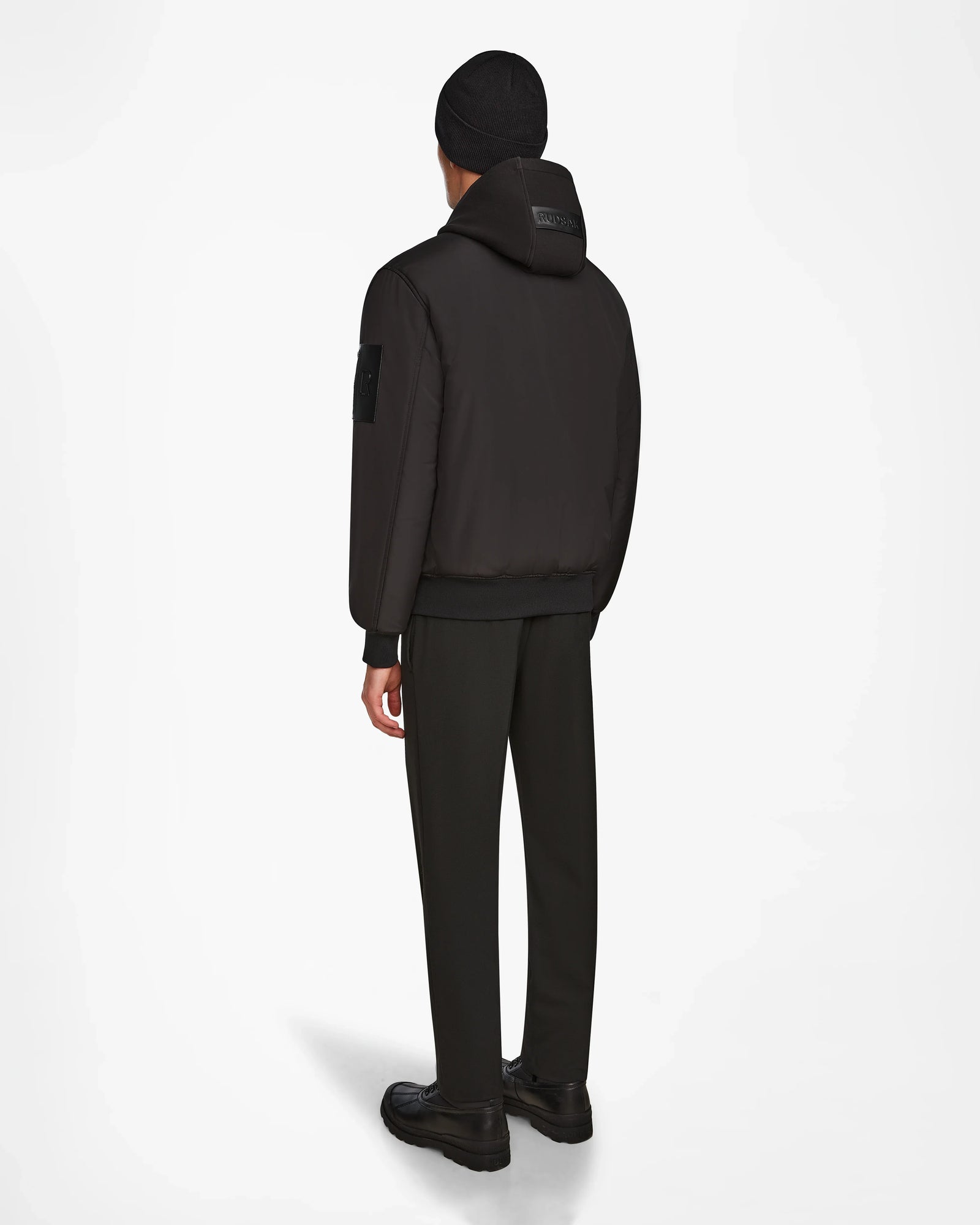 Wearing the Rudsak NEVEN - M124520 Black men’s bomber jacket, a person stands facing away in dark dress pants, black beanie, and black shoes against a white background.