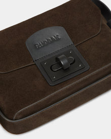 A close-up of the Rudsak OFILIA S - 8324521 Espresso, a dark brown suede crossbody bag with a black leather flap, embossed RUDSAK logo, and a rectangular black metal clasp on the front.