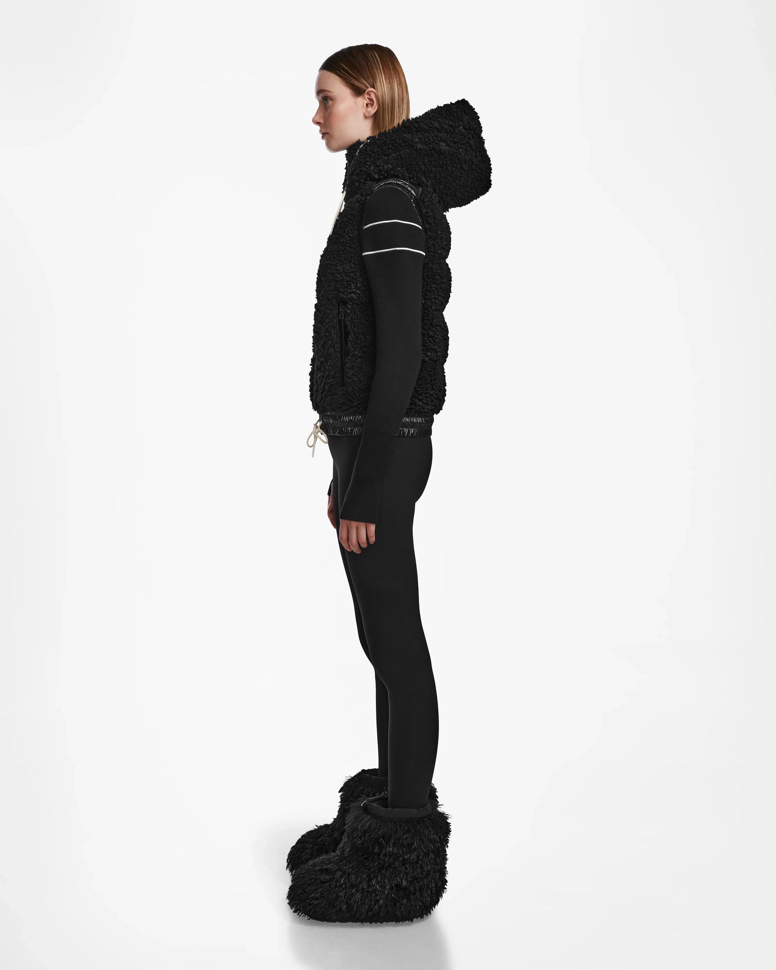 A person stands in profile against a white background, wearing the RAQUEL S - 8124526 Black vest by Rudsak, paired with black fitted pants and oversized black furry boots. Their hair is straight and they have a neutral expression—perfect for winter mountain adventures.