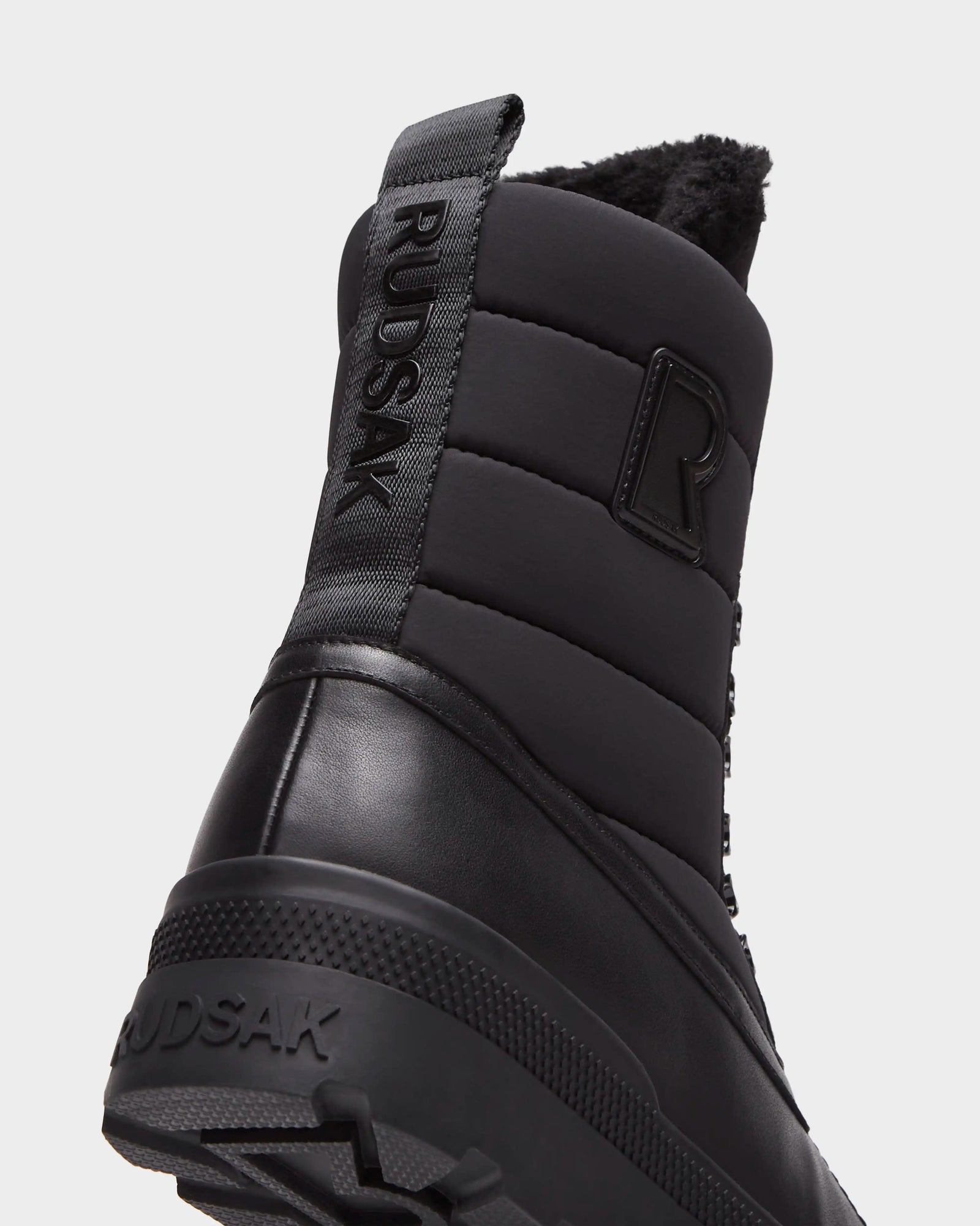 Close-up of the heel of Rudsak’s REAGAN - 6224501 Black men’s winter boots, highlighting a quilted upper, branded pull tab, textured rubber sole, and fuzzy black lining for stylish warmth.