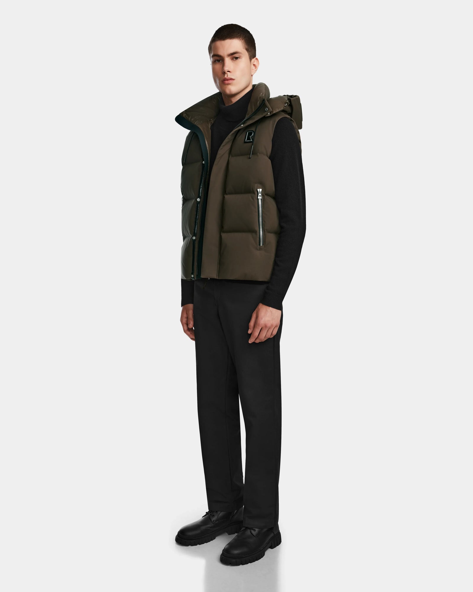A young man faces forward on a white background, wearing the Rudsak REN M - 9123512 Army M weather-resistant dark green puffer vest over a black turtleneck, with black pants and shoes.