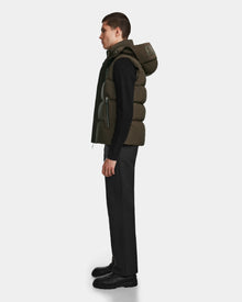 A man stands in profile against a plain background, wearing the Rudsak REN M - 9123512 Army M weather-resistant dark green puffer vest with a hood, paired with a black long-sleeve shirt, black pants, and black shoes.