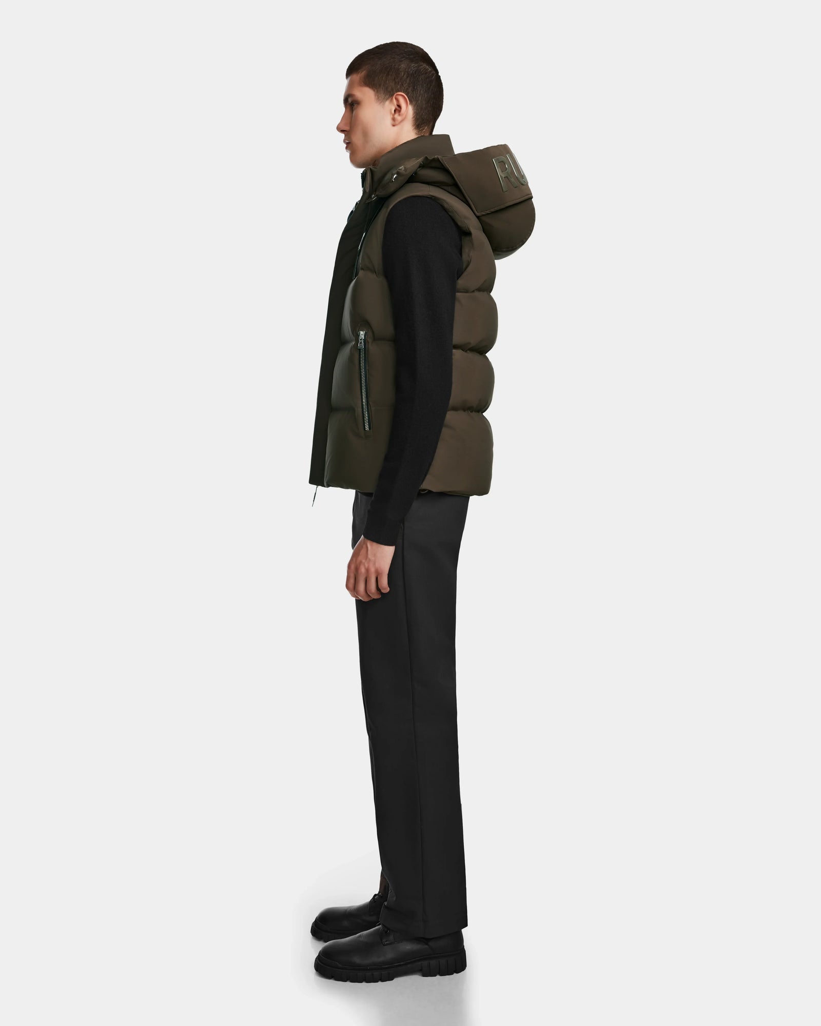 A man stands in profile against a plain background, wearing the Rudsak REN M - 9123512 Army M weather-resistant dark green puffer vest with a hood, paired with a black long-sleeve shirt, black pants, and black shoes.