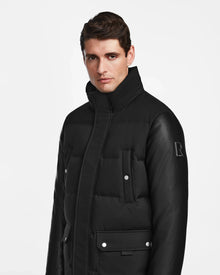 A man with short brown hair wears the Rudsak Ricky R - M123506 Black men's quilted down parka with recycled leather sleeves, a high collar, buttoned pockets, and a subtle R logo on the sleeve, standing against a plain white background.