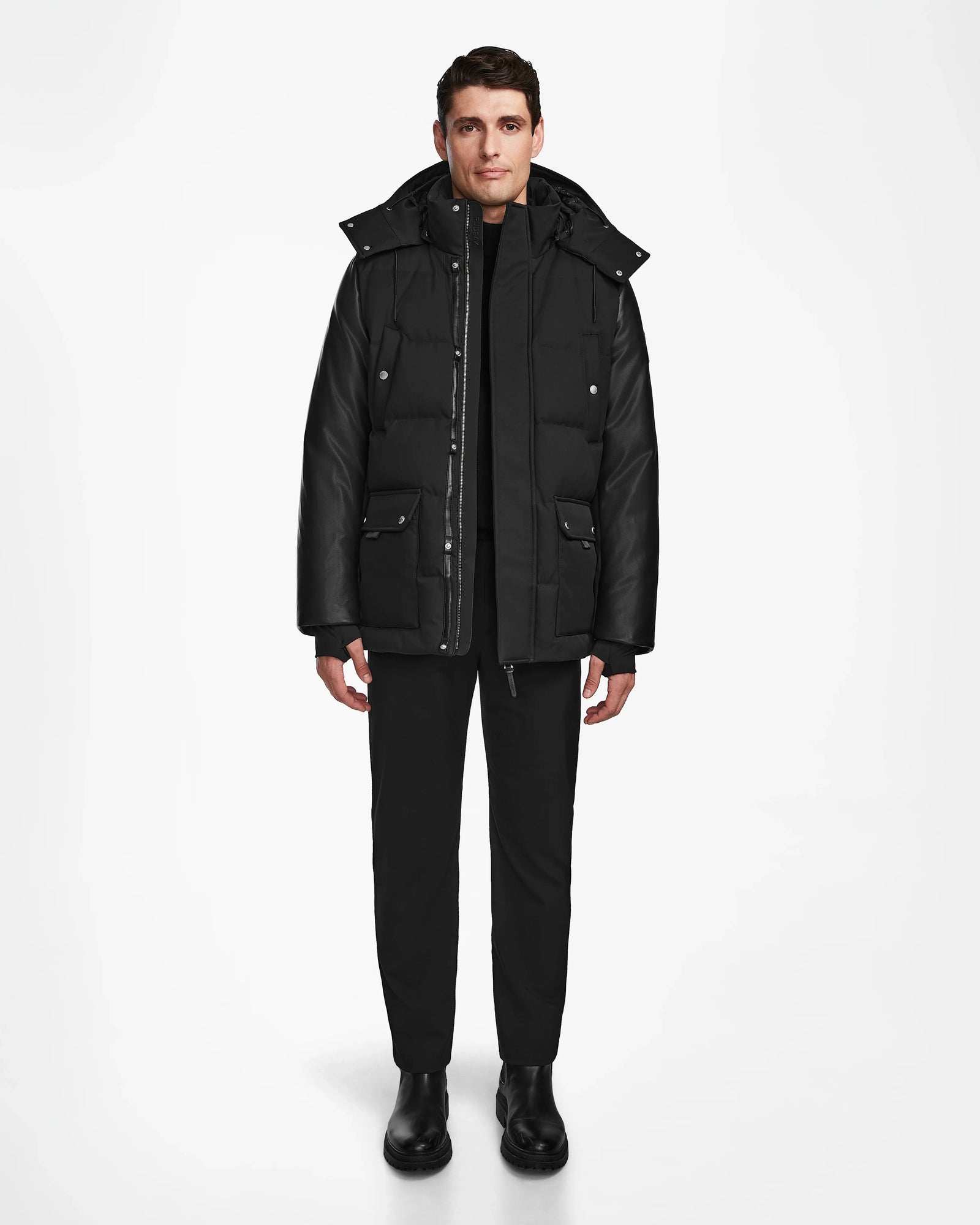 A man stands against a plain white background wearing the Rudsak RICKY R - M123506 Black men's quilted down parka with recycled leather sleeves, paired with black pants and boots. He faces forward, arms relaxed at his sides.
