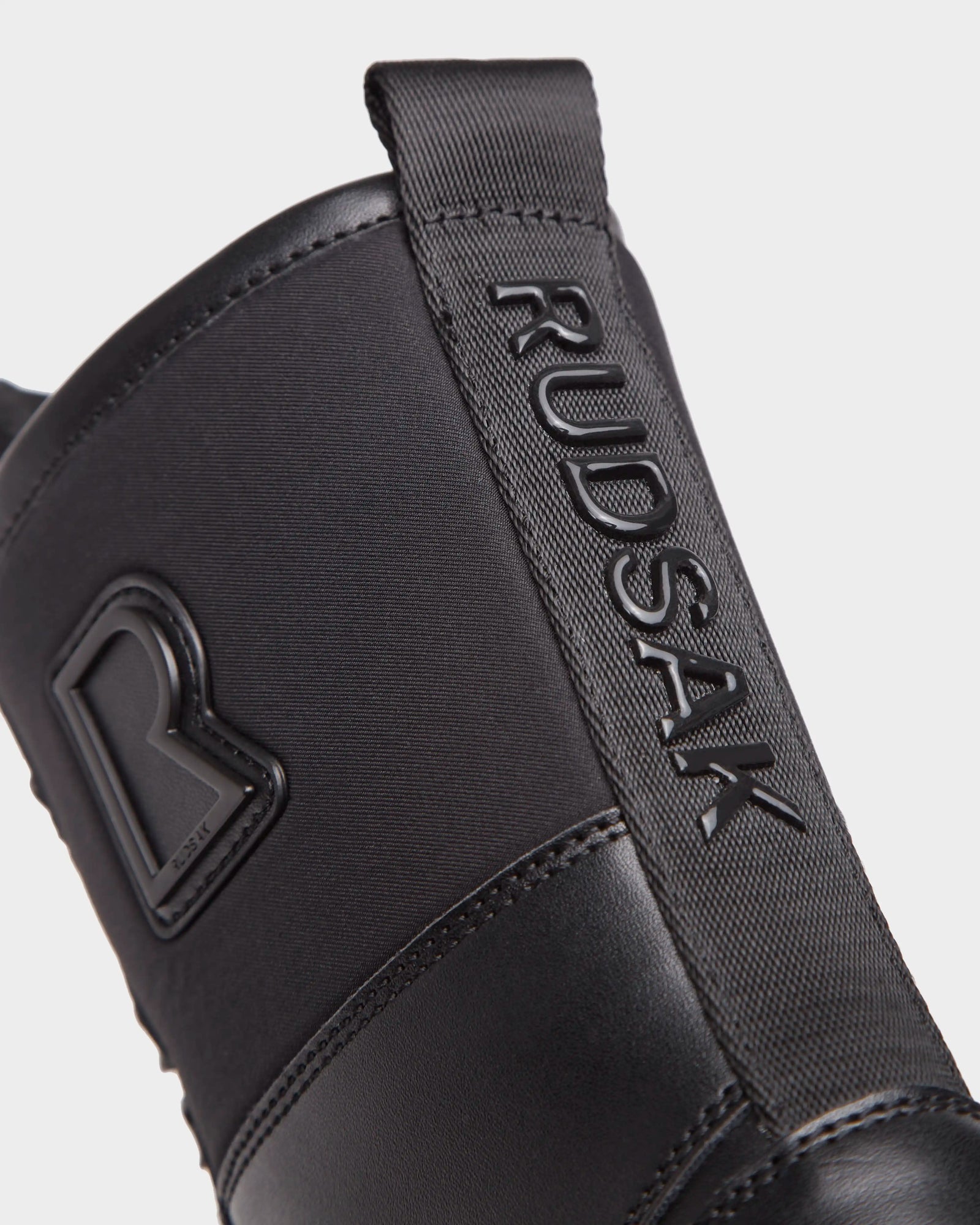 Close-up of the RITA - L222502 Black women's winter boot by Rudsak, with weather-treated leather, "RUDSAK" on a fabric pull tab, raised R logo on the side, and visible stitching details.