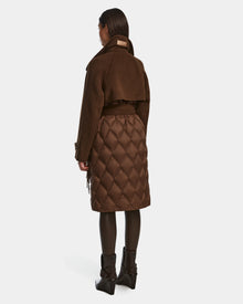 A woman with long, straight hair stands in a neutral pose wearing the Rudsak ZOE - 8124711 Mocha womens quilted down wool trench with a solid upper and quilted lower half, dark tights, and black block-heeled ankle boots.