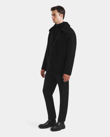 A man stands against a white background wearing the Rudsak AIDEN - 6125558 Black men’s wool-blend coat with removable faux shearling collar, paired with a black turtleneck, pants, and shoes, facing slightly right to display this refined outerwear.
