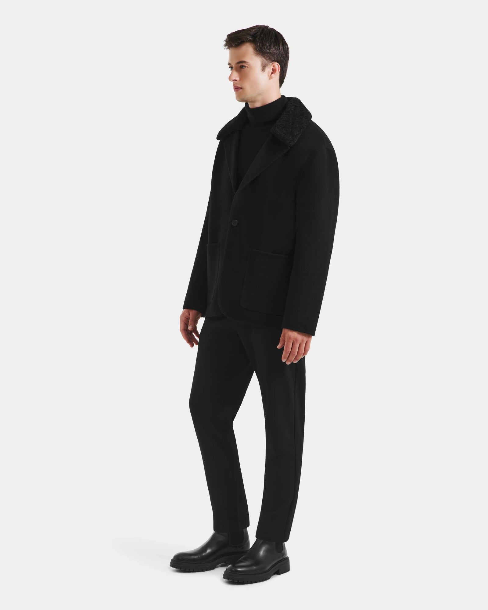 A man stands against a white background wearing the Rudsak AIDEN - 6125558 Black men’s wool-blend coat with removable faux shearling collar, paired with a black turtleneck, pants, and shoes, facing slightly right to display this refined outerwear.