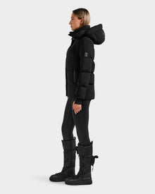 A person in profile wears the Rudsak ALESSANDRA S-M - L125551 Black down puffer jacket with hood, styled with black leggings and winter boots, standing against a plain light background.